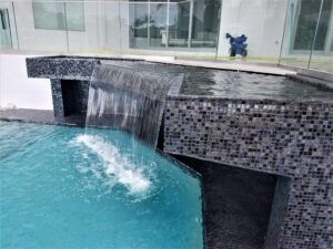 A raised spa clad in dark mosaic tile spills water over an edge into the main pool below.