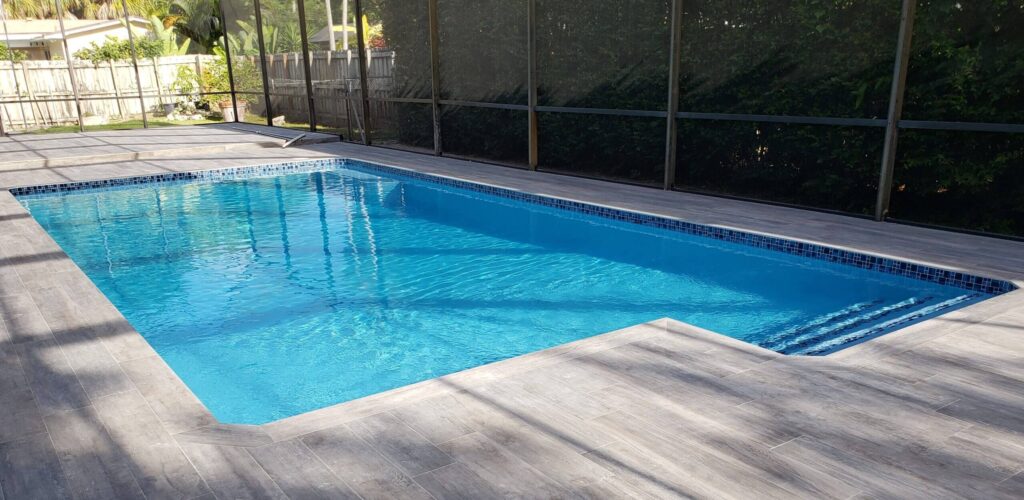 A rectangular pool with a tiled waterline and wood-look tile decking inside a screened enclosure.