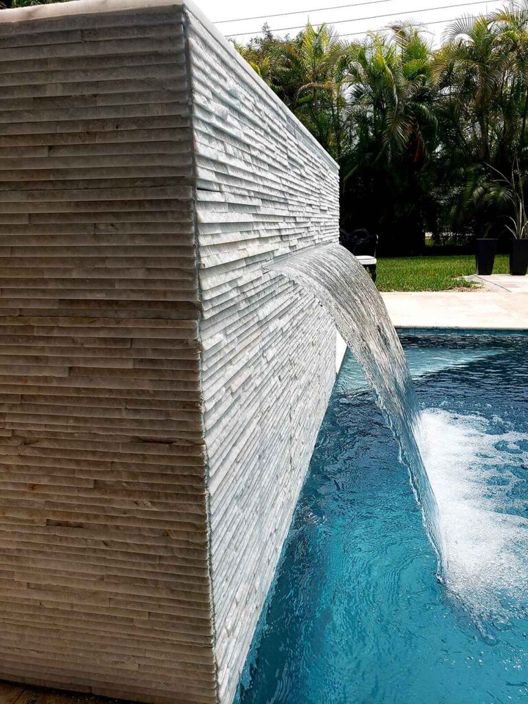 A stacked-stone water feature wall pours a sheet of water into a pool with a paved patio beyond.