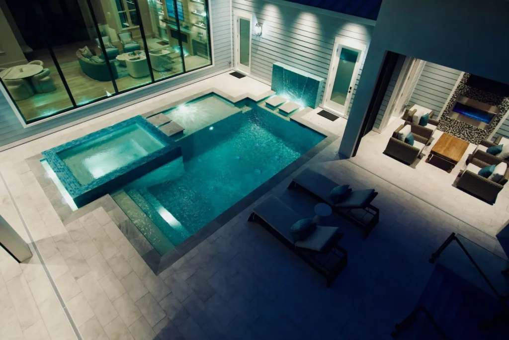 An elevated night view of a sleek rectangular pool with a raised glass-tiled spa and water feature wall, nestled in a modern courtyard between glass walls and an outdoor lounge.