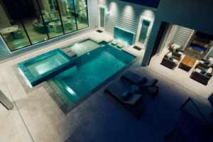 An elevated night view of a sleek rectangular pool with a raised glass-tiled spa and water feature wall, nestled in a modern courtyard between glass walls and an outdoor lounge.