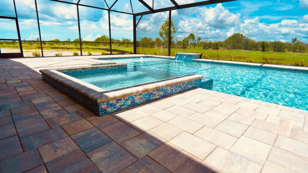 A raised spa with glass tile walls spills water into a screened pool overlooking open fields.