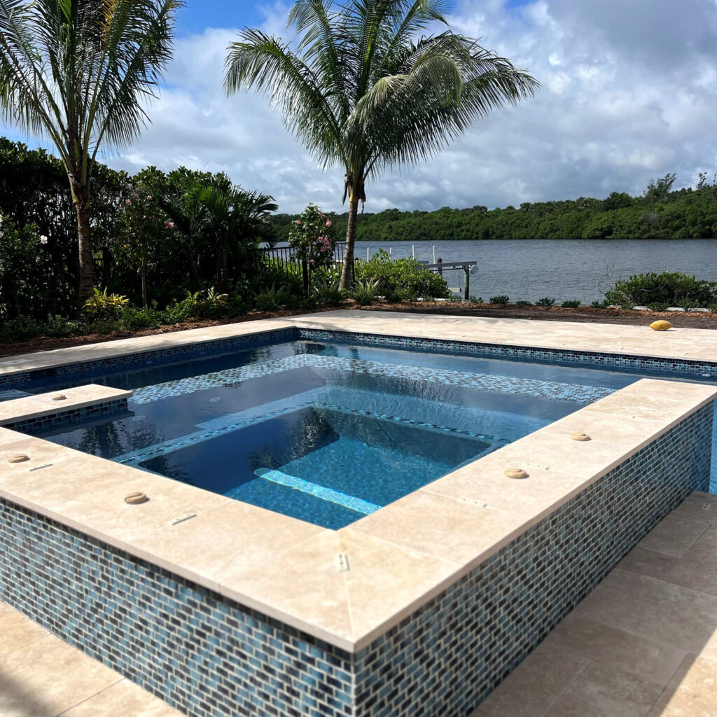 A raised spa with blue mosaic tile and travertine coping sits beside a waterway lined with palm trees.