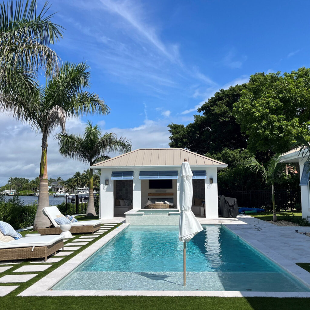 A long rectangular pool with a raised spa in front of a waterfront cabana with palm trees.