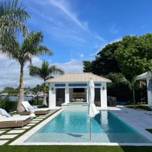 A long rectangular pool with a raised spa in front of a waterfront cabana with palm trees.