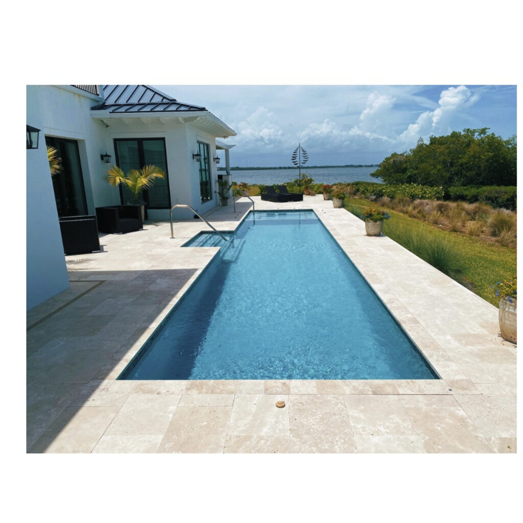 A lap pool with a small spa at one end sits on a travertine patio overlooking a waterfront view.
