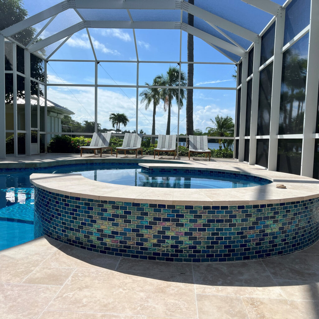 A raised spa clad in shimmering blue glass mosaic tile sits beside a pool under a screened lanai with a canal view.