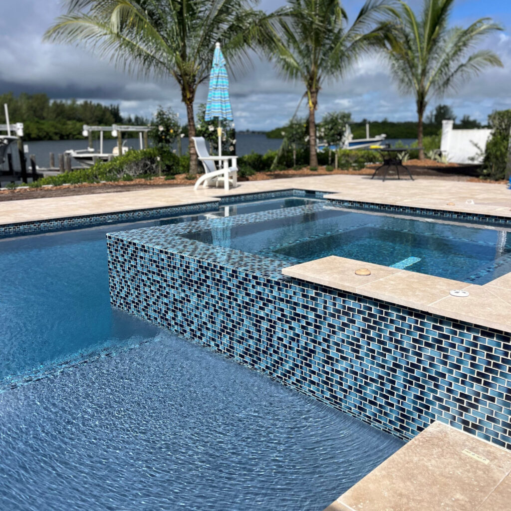 A raised spa clad in blue glass mosaic tile spills into a pool with a canal and palm trees behind.