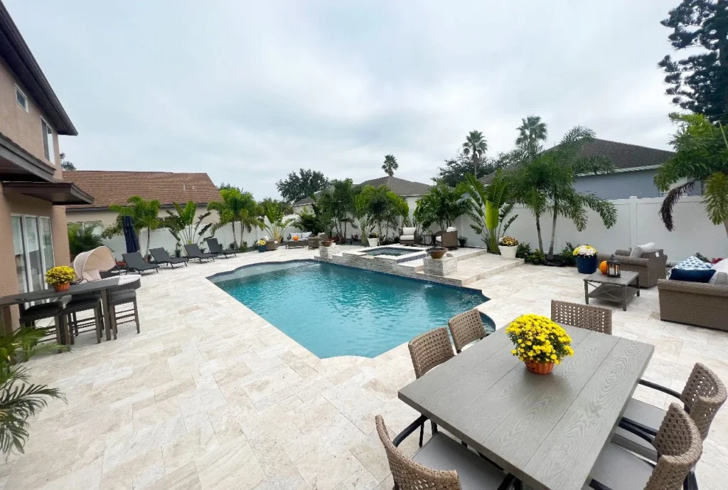 A freeform backyard pool with an attached raised spa surrounded by a large travertine patio and outdoor furniture.