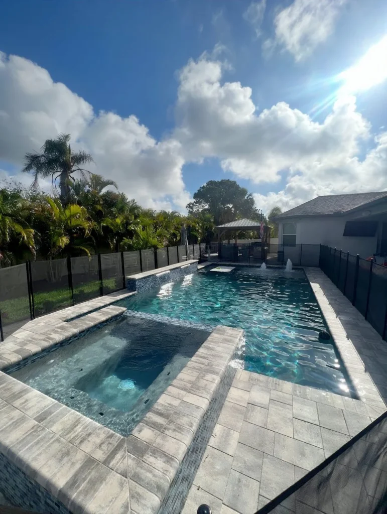 Rectangular backyard pool with raised spa and paver deck under a sunny sky, enclosed by a black safety fence.