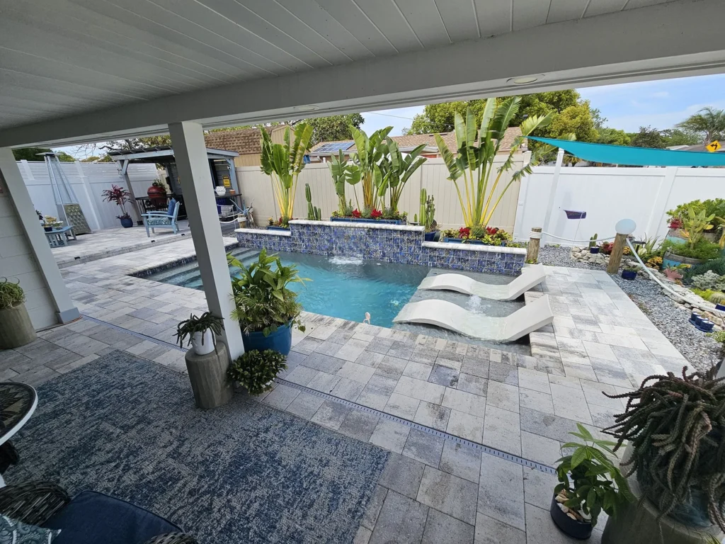 A small backyard pool with a raised spa, tiled water feature wall, and submerged loungers under a covered patio.