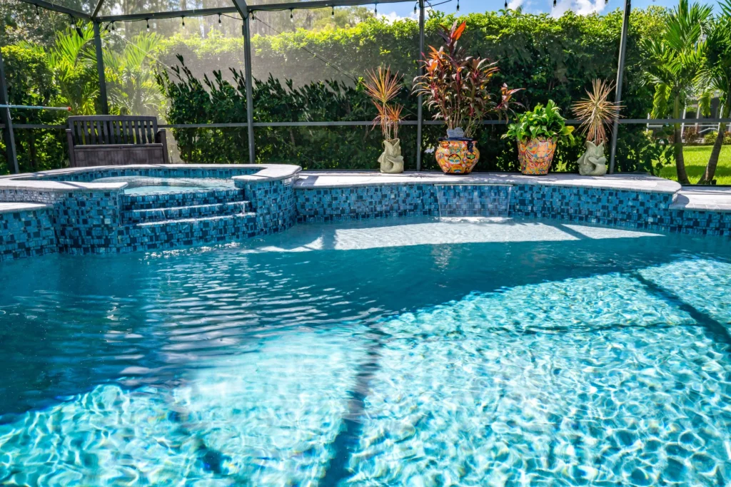 A blue mosaic-tiled freeform pool with a raised spa and small waterfall steps under a screened enclosure.