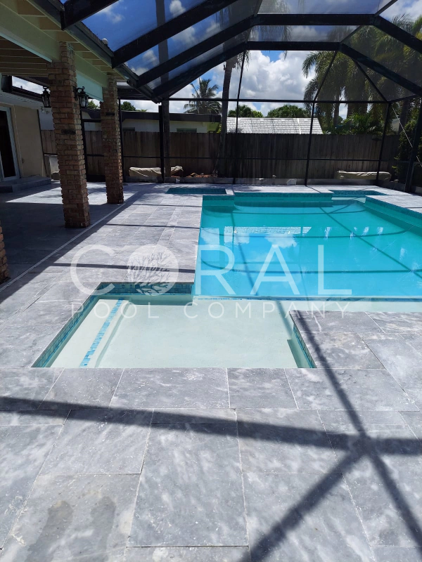 Rectangular pool with sun shelf and spa under a screened lanai, surrounded by gray travertine pavers.