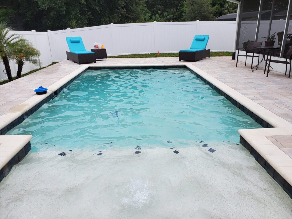 A rectangular backyard pool with a shallow beach-style entry, travertine deck, and two teal loungers beside a white vinyl fence.