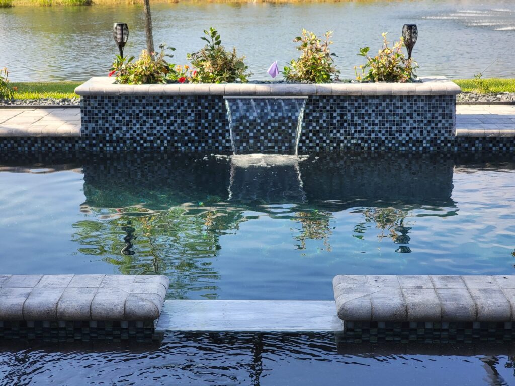 A tiled raised wall with a small waterfall spills into a pool overlooking a lake.