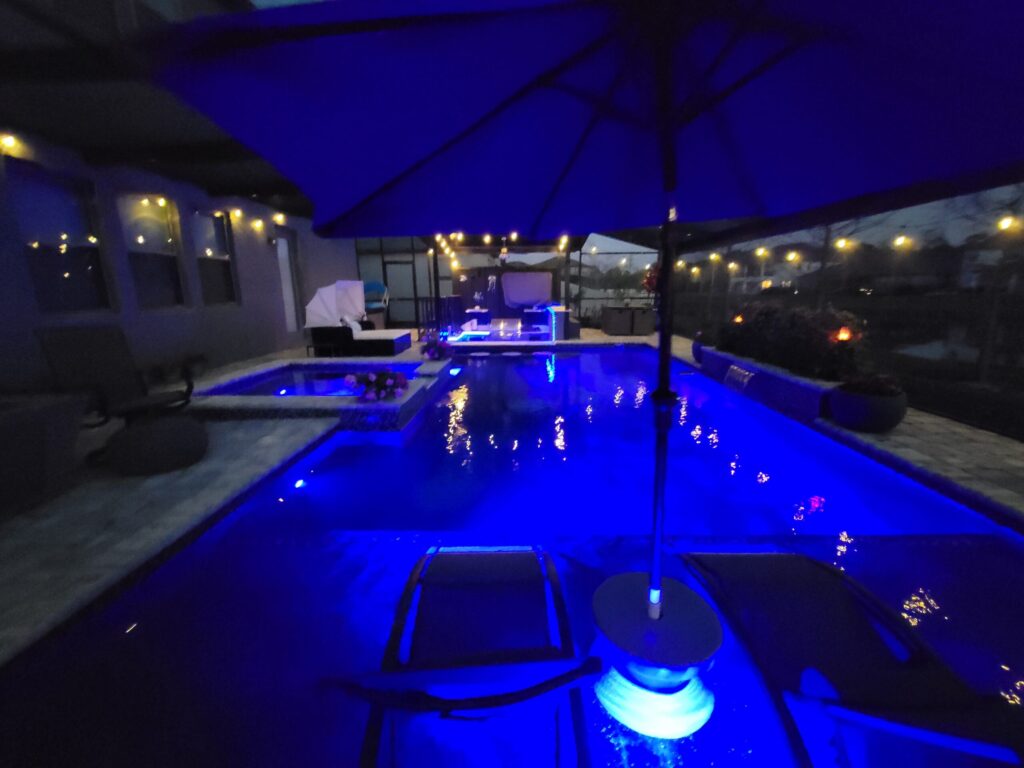A backyard pool lit with vivid blue LED lighting at night, with an attached raised spa and umbrella-shaded loungers.