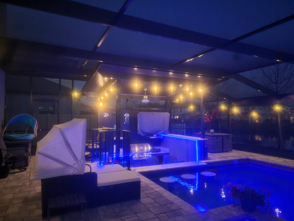 Nighttime patio with string lights, outdoor kitchen, and a corner of a blue-lit pool under a screen enclosure.