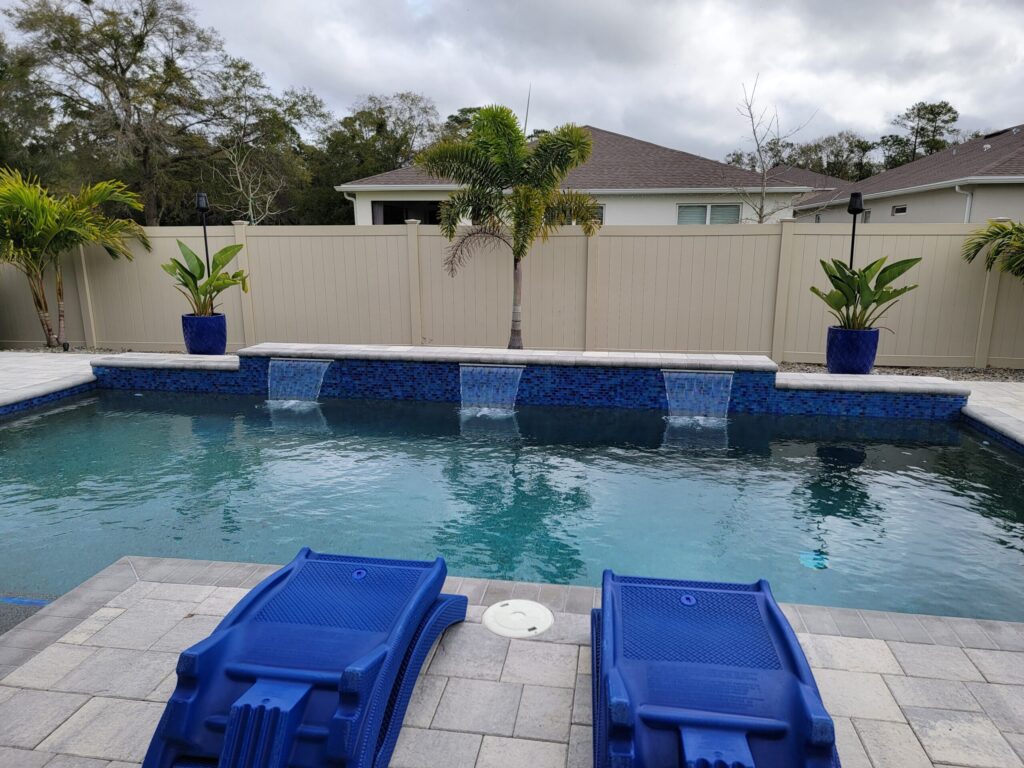 A rectangular backyard pool with three scupper water features flowing from a blue tile raised wall, framed by a beige privacy fence and palm trees.