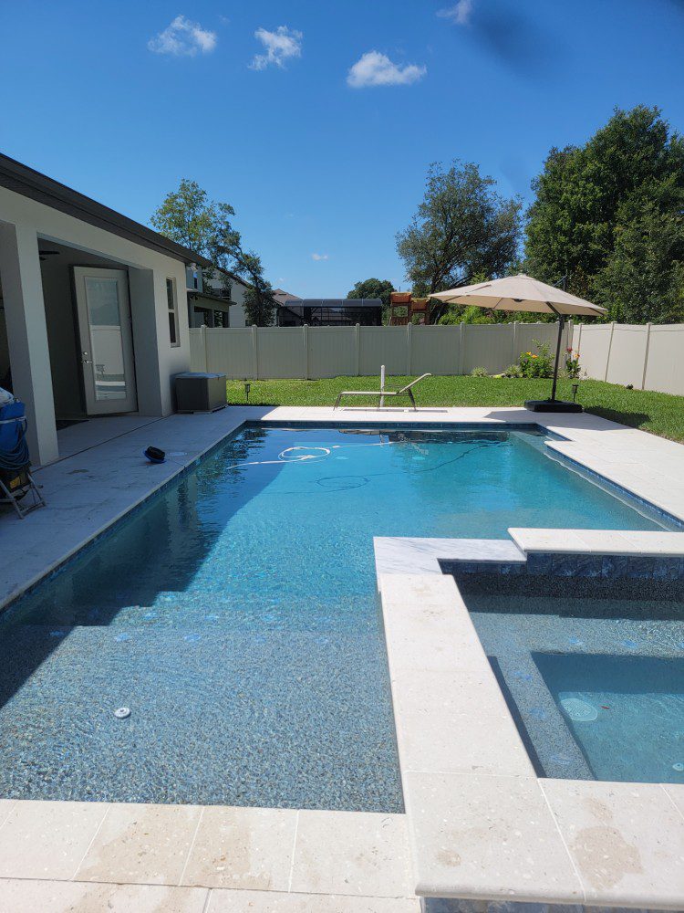 A rectangular backyard pool with an attached raised spa and travertine deck under a clear blue sky.