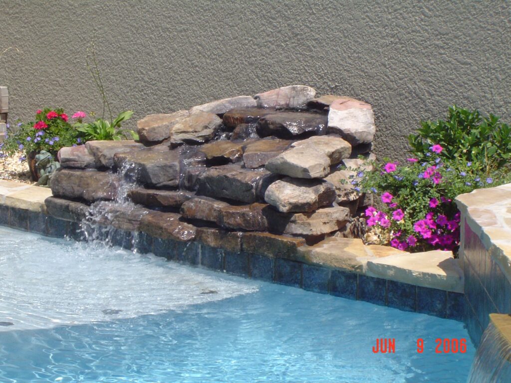 A stacked-stone waterfall feature cascades into a pool corner surrounded by flowering plants.