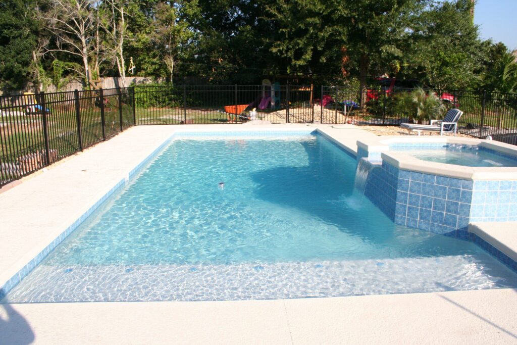 A rectangular vinyl-liner pool with a raised tiled spa spilling into it, enclosed by a black metal fence in a backyard with a play area.