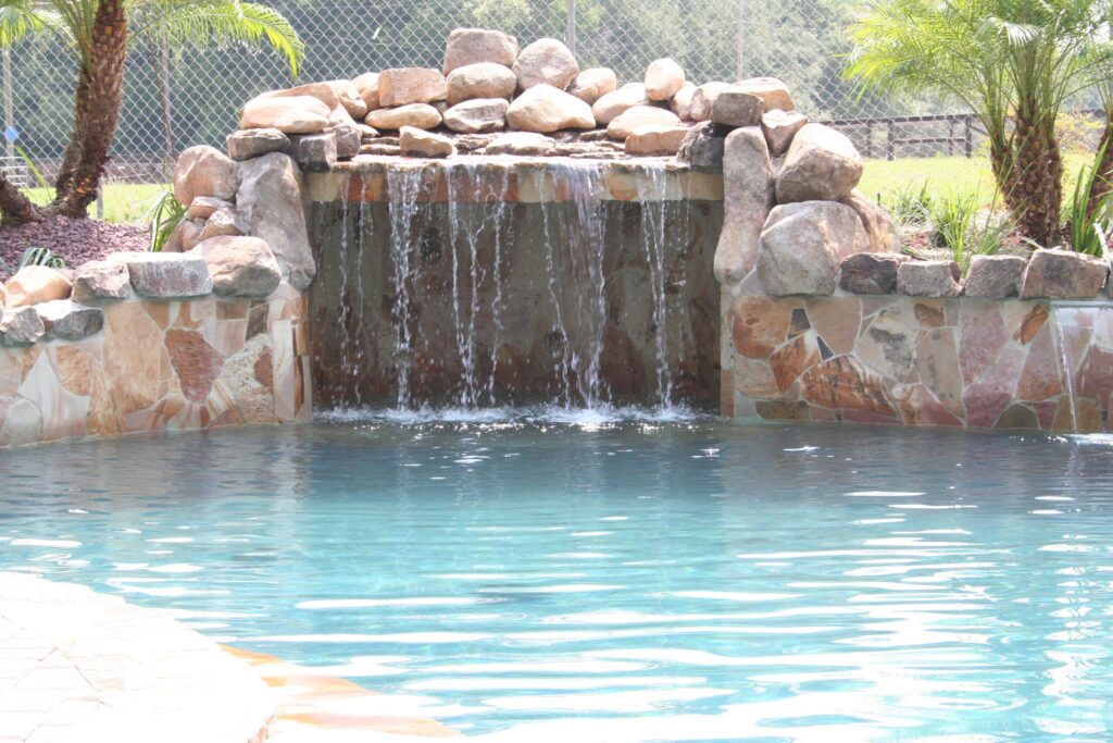 A stacked-boulder waterfall cascades over a stone wall into a backyard pool.