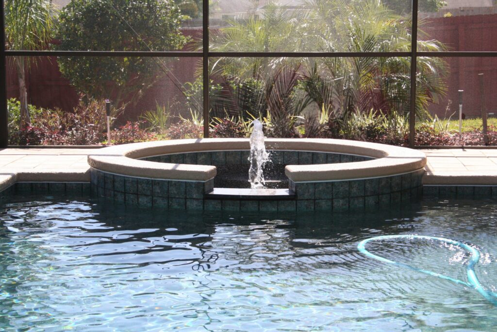 A raised tiled fountain feature bubbles water into a screened pool with a pool net floating nearby.