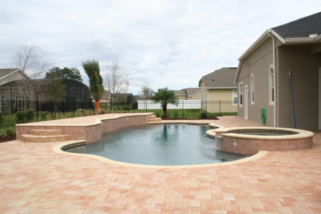 A freeform backyard pool with a raised spa spilling into it, surrounded by a paver patio under an overcast sky.