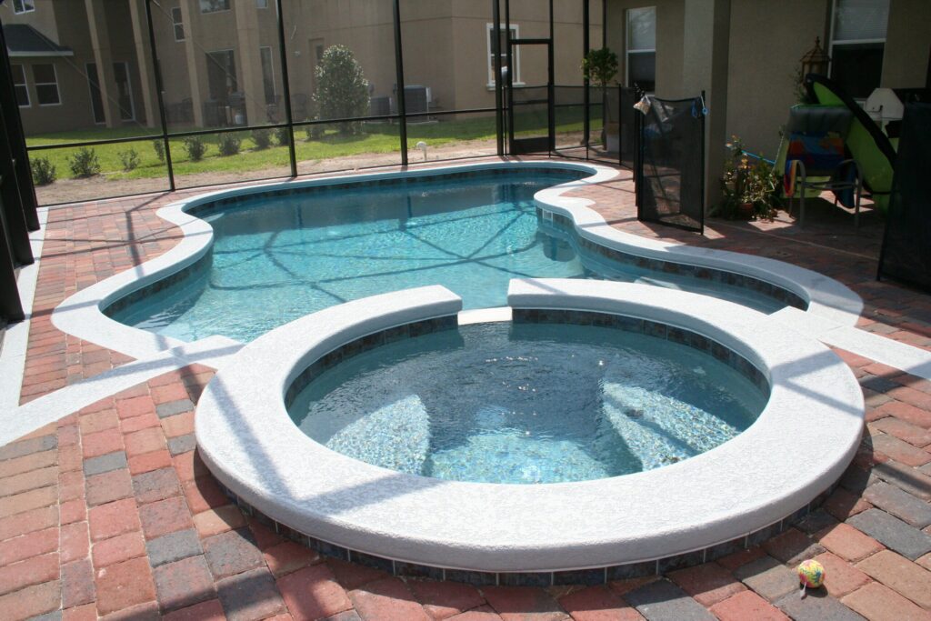 A freeform pool with a raised round spa inside a screened lanai, patio with a child safety fence and pool toys.