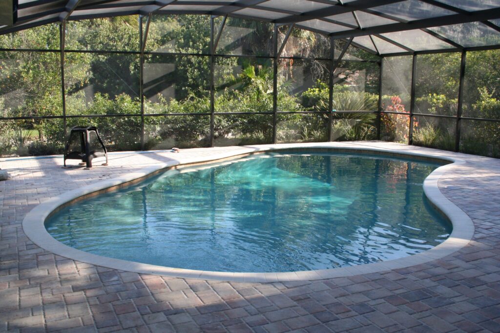 A kidney-shaped pool with light plaster finish under a screened enclosure surrounded by lush greenery.