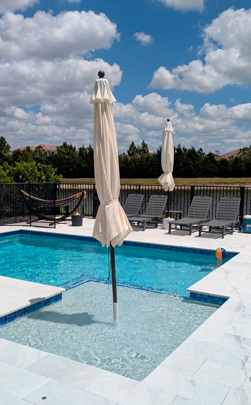 A rectangular backyard pool with a shallow sun shelf and umbrella, lounge chairs and a hammock on a paver deck.