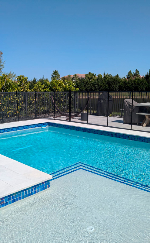 Rectangular backyard pool with blue tile waterline and a broad shallow entry step, framed by a black safety fence and patio furniture.