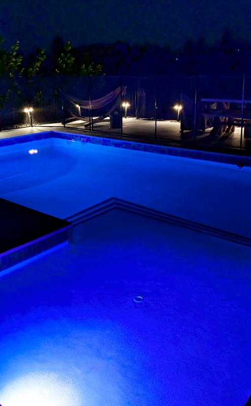 A rectangular backyard pool glowing blue at night with a tanning ledge and glass fence.