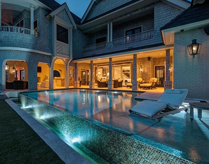 A glass-tiled infinity-edge pool glows at night beside a large shingle-style home with a covered lanai.