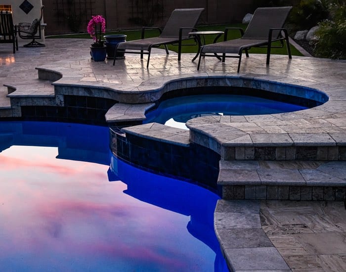 A raised spa with dark tile spills toward a freeform pool on a travertine paver patio at dusk.