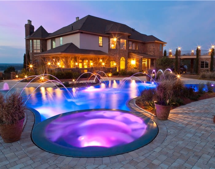 A raised spa lit purple sits in front of a pool with arcing water jets before a large estate home at dusk.