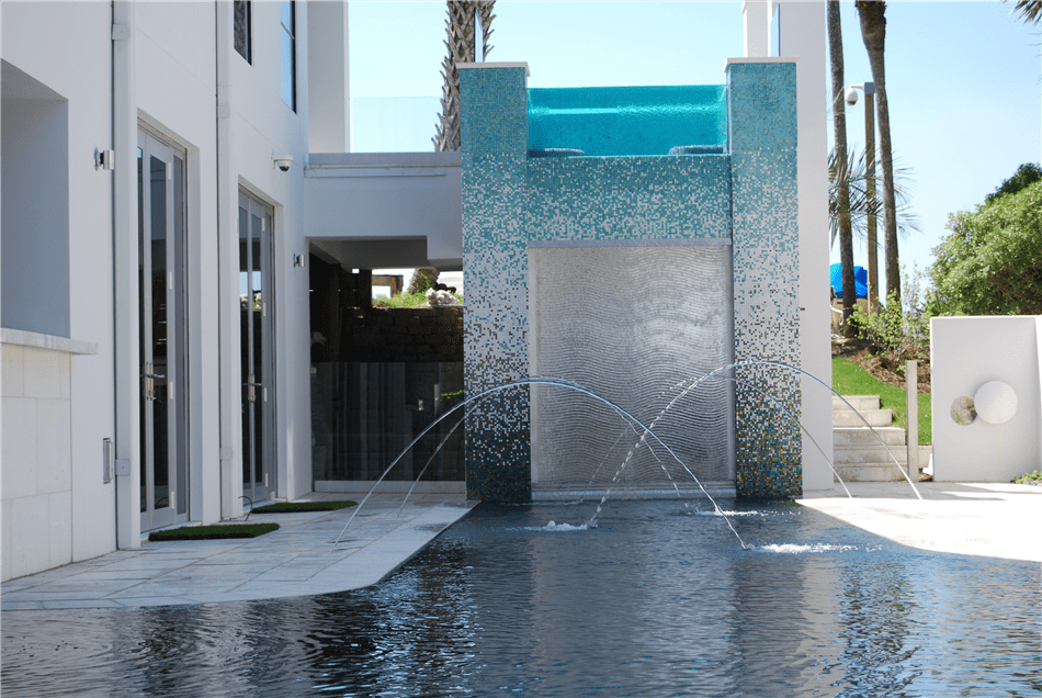 Modern mosaic-tiled water feature wall with jets arcing into a dark reflective pool beside a contemporary home.