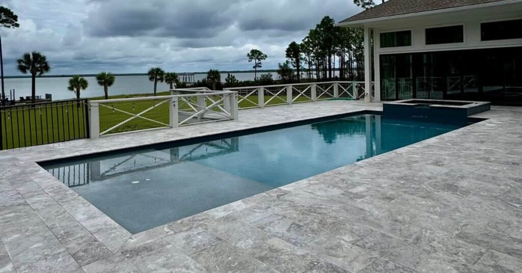 A rectangular gunite pool with a raised spa sits on a travertine patio overlooking a wide waterfront view under a cloudy sky.