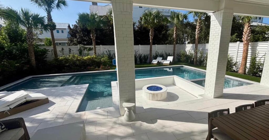 A rectangular backyard pool with a small L-shaped extension and a round fire pit on the stone patio, framed by palm trees and a white privacy fence, viewed from under a covered lanai.