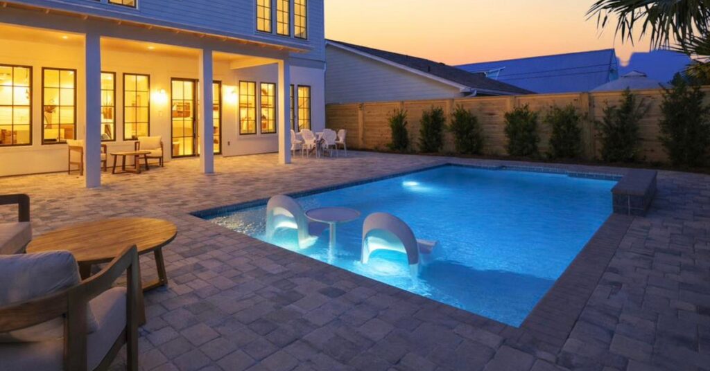 A rectangular backyard pool glowing blue at dusk with in-water table and chairs, surrounded by a paver patio and wood fence.