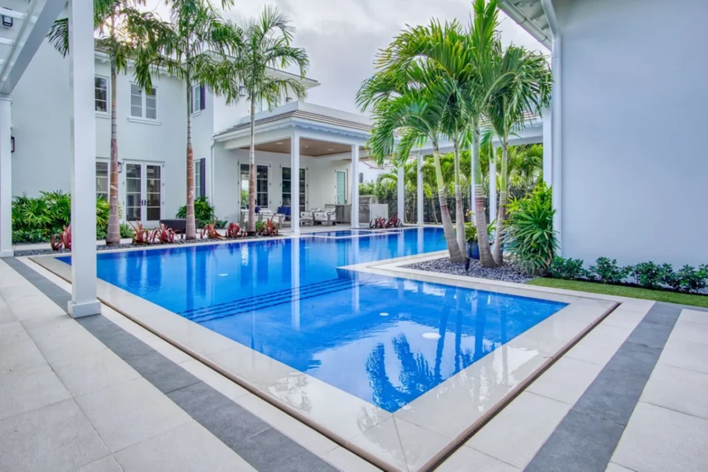 An L-shaped pool with a sun shelf runs along a modern white two-story home framed by tall palm trees.