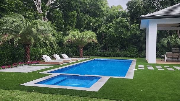 A rectangular backyard pool with an attached spa, surrounded by turf lawn, loungers, and tropical landscaping.