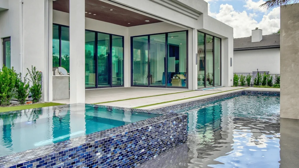 A long lap pool with a mosaic-tiled overflow edge runs alongside a modern home's glass-walled terrace.