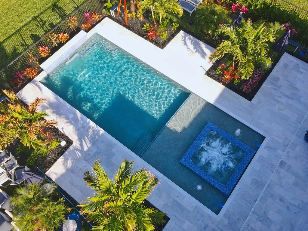 Aerial view of a rectangular backyard pool with a raised square spa, tanning ledge, and travertine deck surrounded by tropical landscaping.