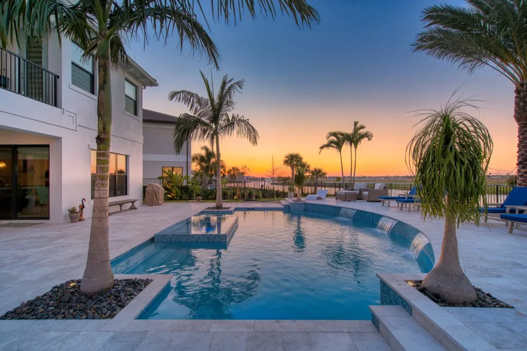 A freeform pool with a raised spa and spillover edges glows at sunset beside a waterfront home lined with palm trees.