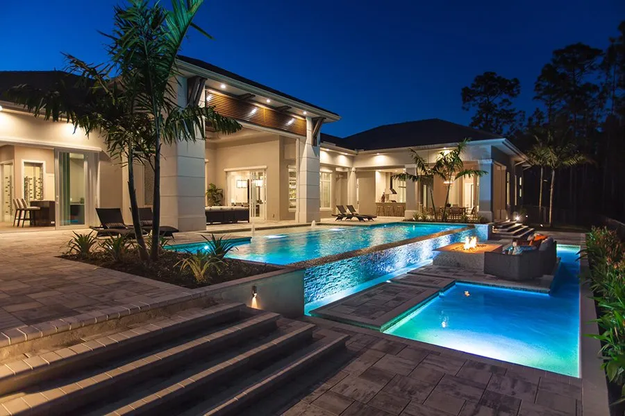An elevated infinity-edge pool spills into a lower pool beside a lit fire pit at night.