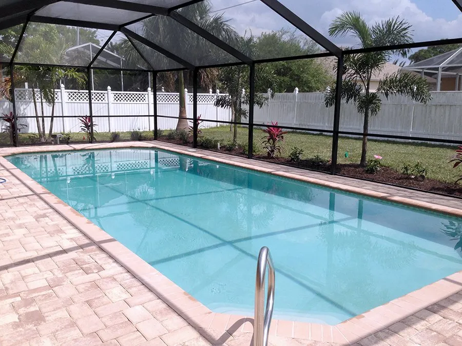 A rectangular pool with turquoise water inside a screened enclosure with a paver deck and white vinyl fence backdrop.