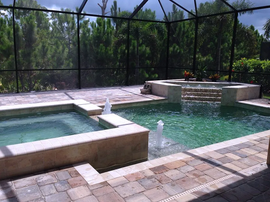 A geometric pool with a raised spa and tiled waterfall steps sits under a screened lanai surrounded by pine trees.