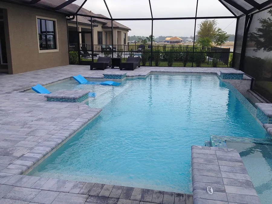 An L-shaped pool with a paver deck and two floating loungers under a screened enclosure.