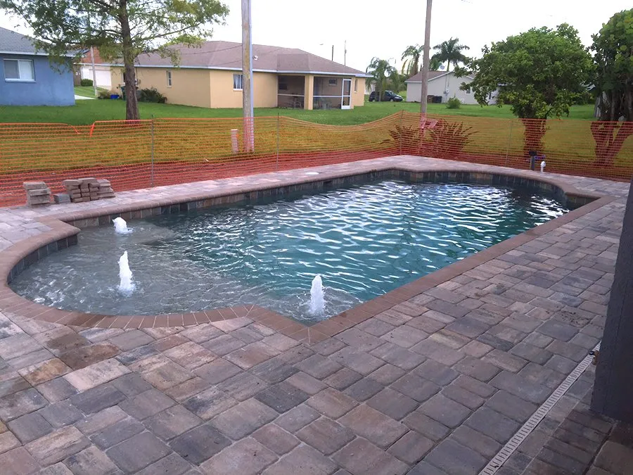 A freeform pool with paver decking and bubbler jets, construction fencing still visible in the yard.